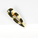 Barrette en résine avec un morceau de chute de pagne wax blanc et noir parsemé de fines paillettes dorées. Barette avec un bon maintien grâce à une pince alligator dorée