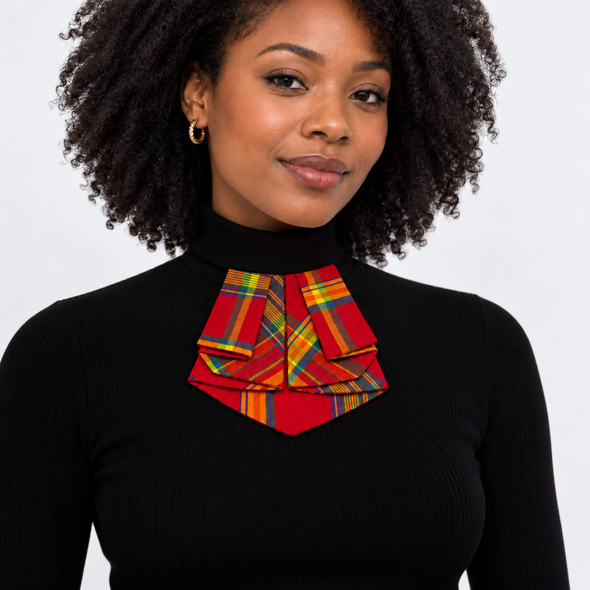 Femme noire aux cheveux naturels portant une cravate courte style jabot en tissu Madras rouge à carreaux multicolores sur un col roulé noir.